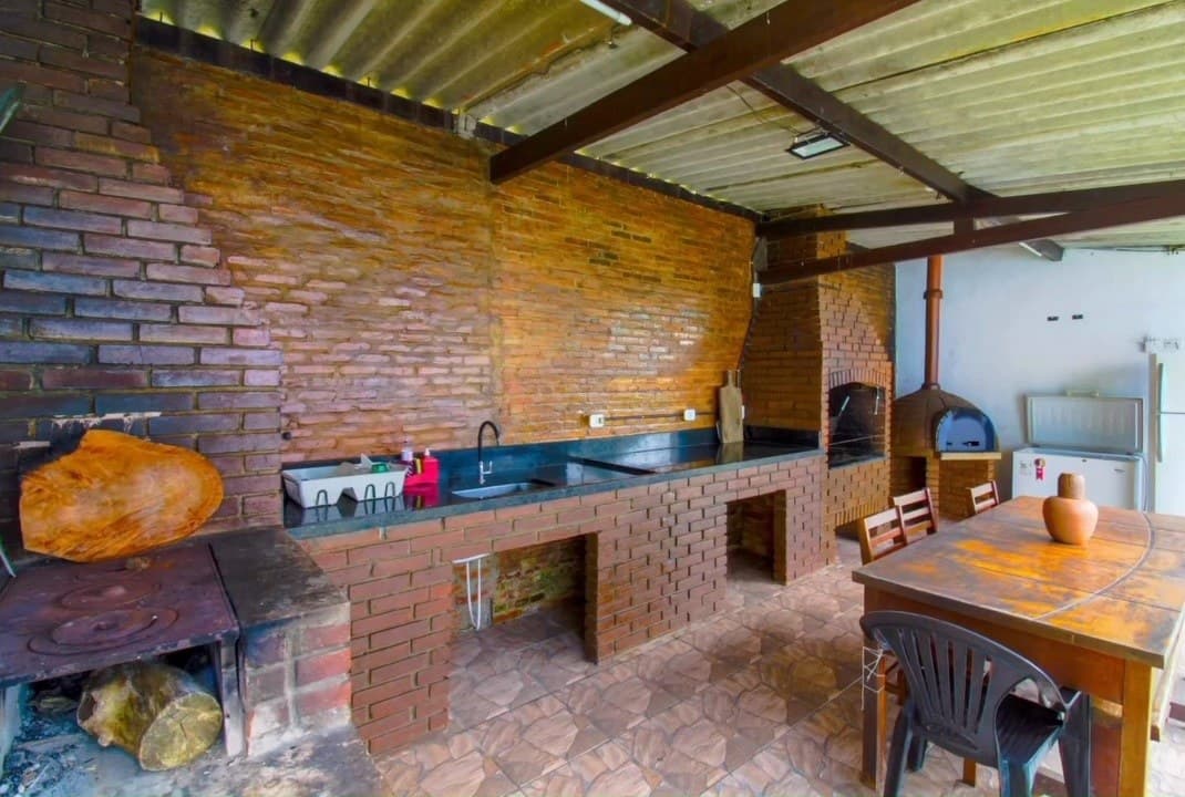 Área gourmet coberta, com parede de tijolos, pia, churrasqueira, forno de pizza e mesa