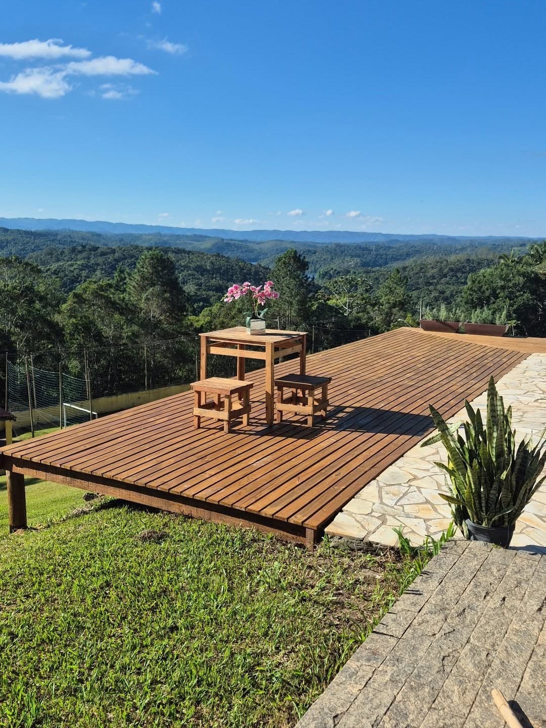 Deck de madeira com mesa e bancos à beira da colina, com a Mata Atlântica e as serras ao fundo