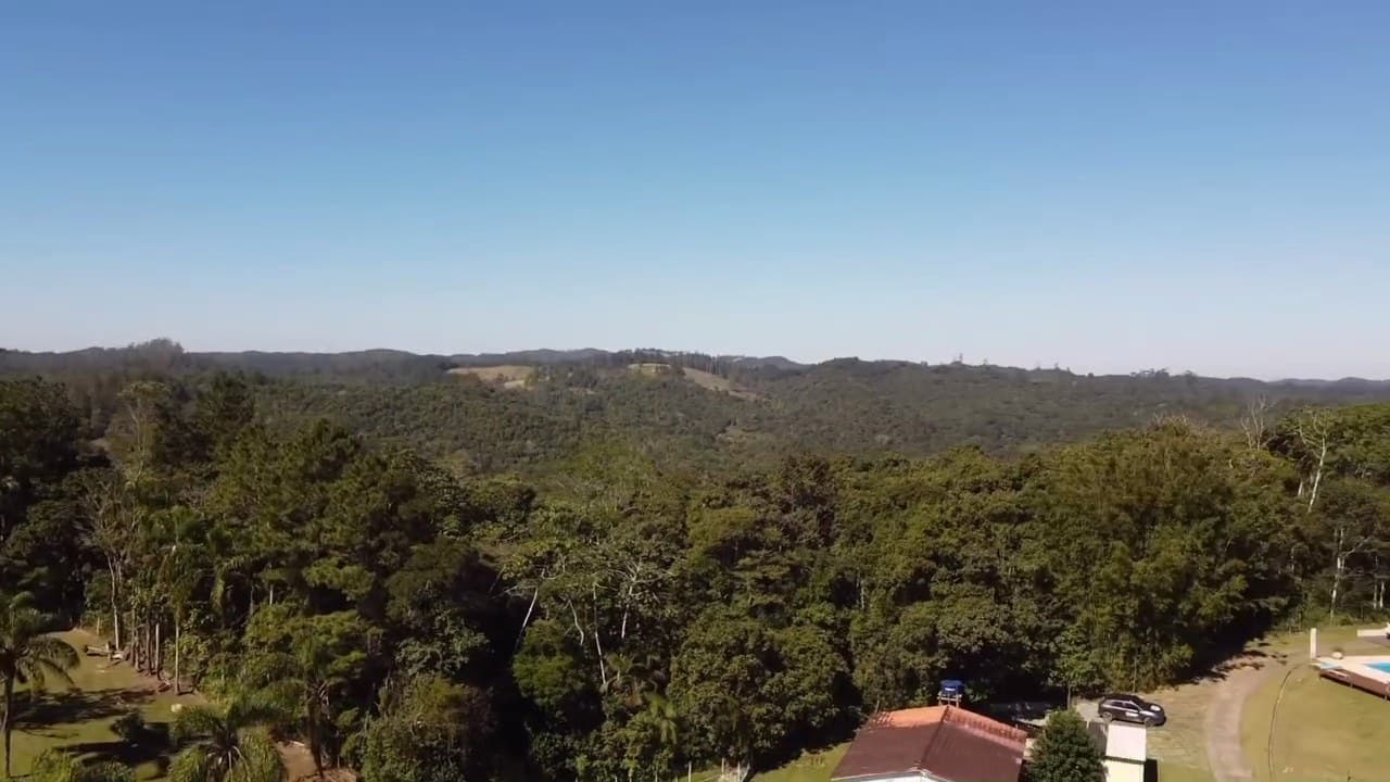 Vista aérea da chácara no alto da colina, com a casa branca, a piscina e o deck cercados de mata