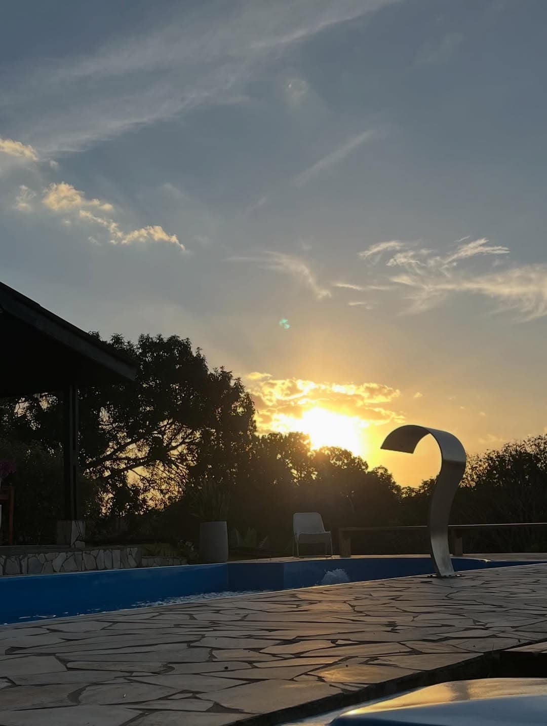 Pôr do sol atrás das árvores, com a piscina e o piso de pedra dourados